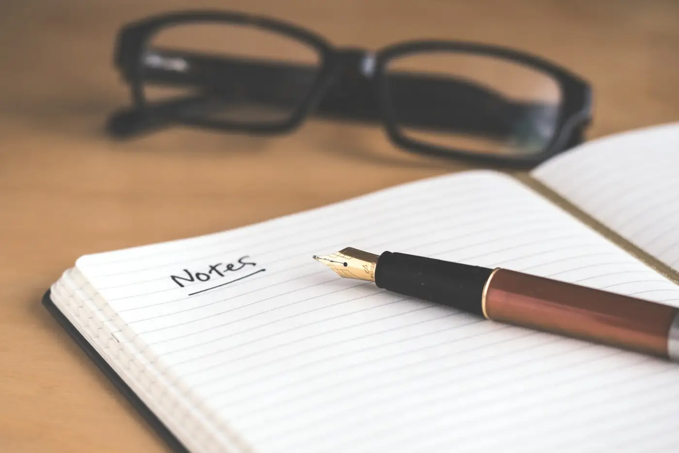 An open notebook with a fountain pen and a pair of glasses on a wooden desk
